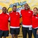 Four people stand smiling in front of a bright yellow backdrop with various sponsor logos. They all wear matching red and white athletic shirts with black shorts. The backdrop features an Ocean event logo prominently.