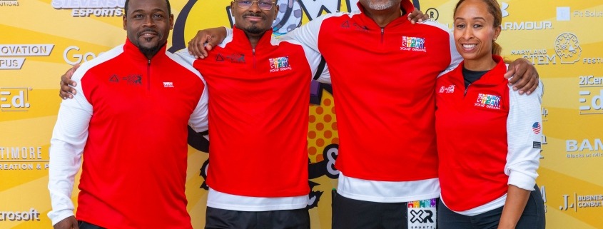 Four people stand smiling in front of a bright yellow backdrop with various sponsor logos. They all wear matching red and white athletic shirts with black shorts. The backdrop features an Ocean event logo prominently.