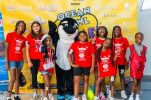 A group of smiling children in red shirts pose with a person in an Orca whale costume in front of a bright yellow Ocean Bowl Live backdrop. Some children make peace signs and all look happy.