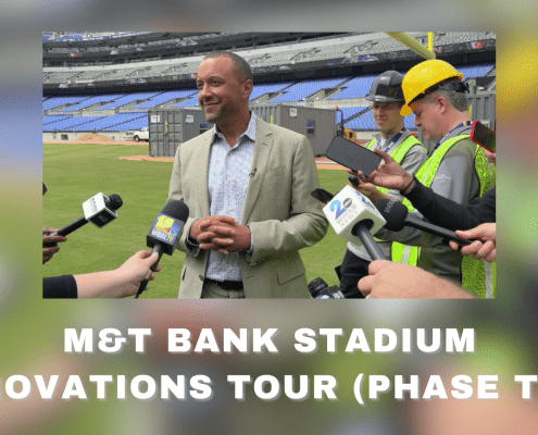 A man in a suit speaks to reporters with microphones at M&T Bank Stadium during renovations, while workers in safety vests and helmets stand nearby. Text reads: M&T Bank Stadium Renovations Tour (Phase Two) and Bowie TV.