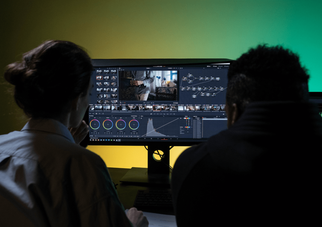 Two people sit facing a large computer monitor displaying video editing software and color grading tools, with a green and yellow gradient background behind them.