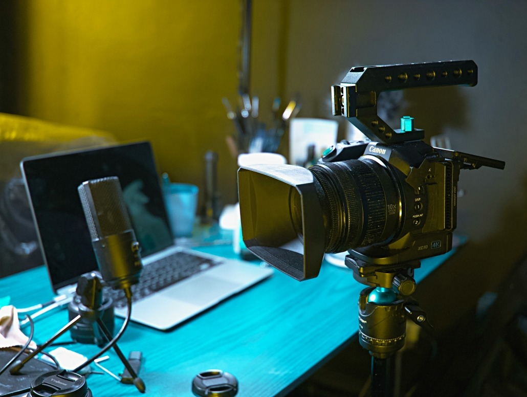 A professional camera on a tripod is set up on a blue desk next to a microphone and an open laptop, with various other small items and blurred background objects visible.