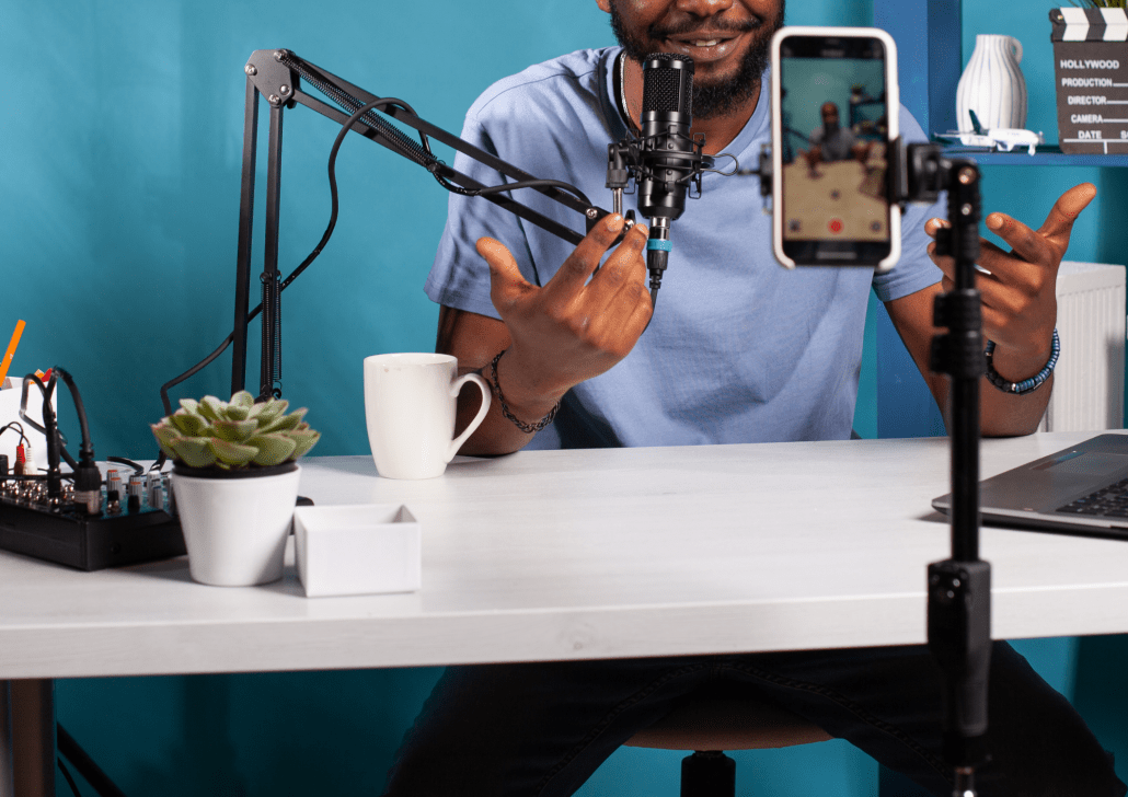 A man sits at a desk recording a podcast or video, speaking into a microphone. He gestures with his hands while a smartphone on a tripod films him. A laptop, coffee mug, plant, and audio equipment are on the desk.